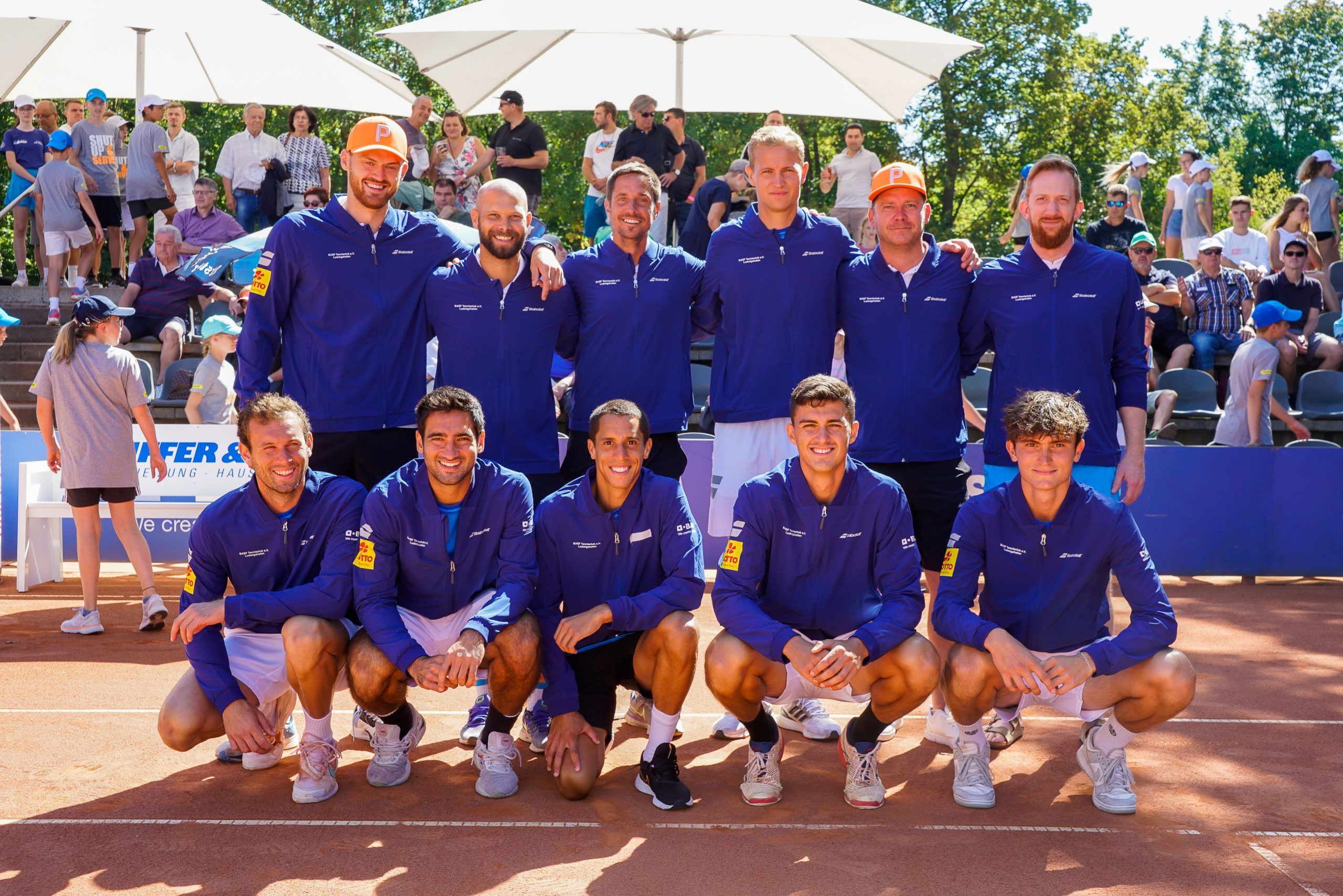 Tennismannschaft posiert auf Sandplatz vor Zuschauern