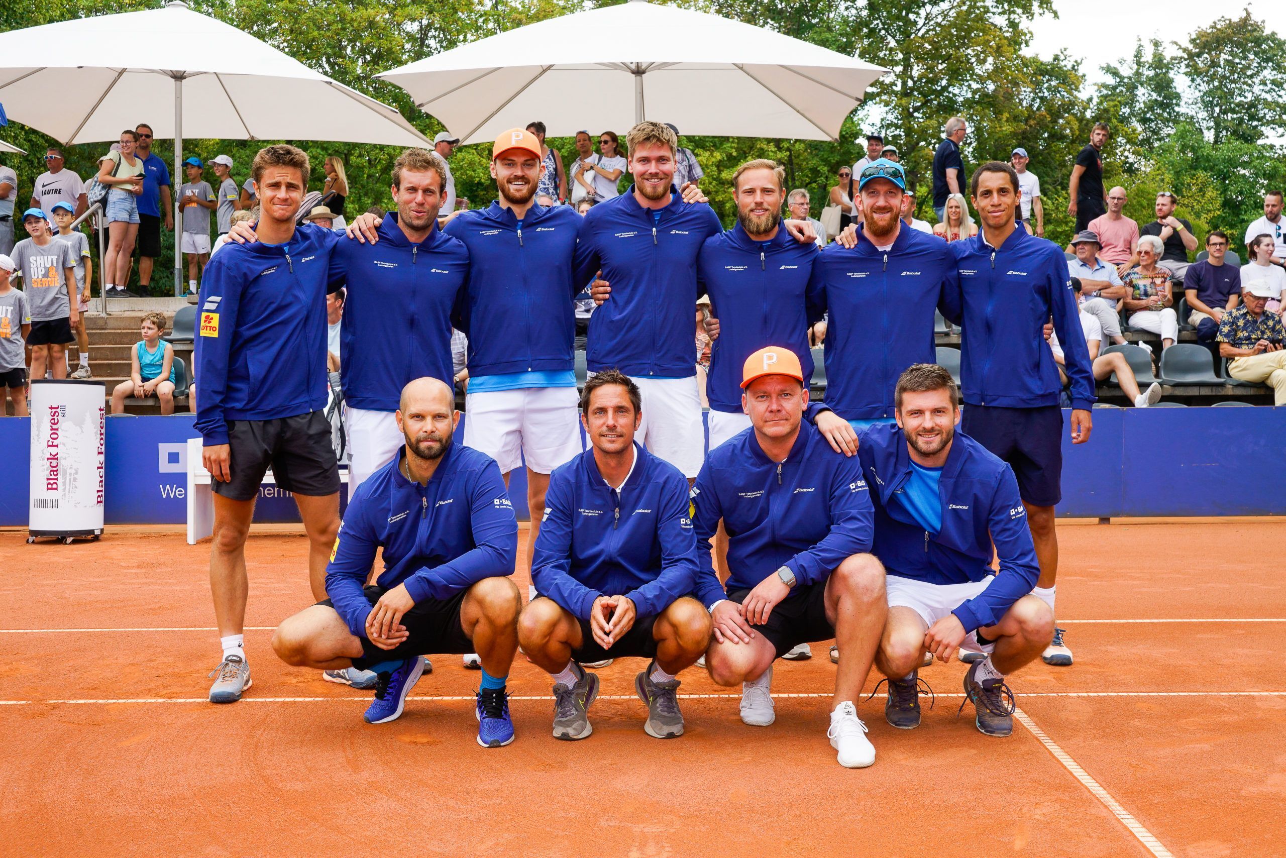 Tennismannschaft posiert auf Sandplatz vor Zuschauern