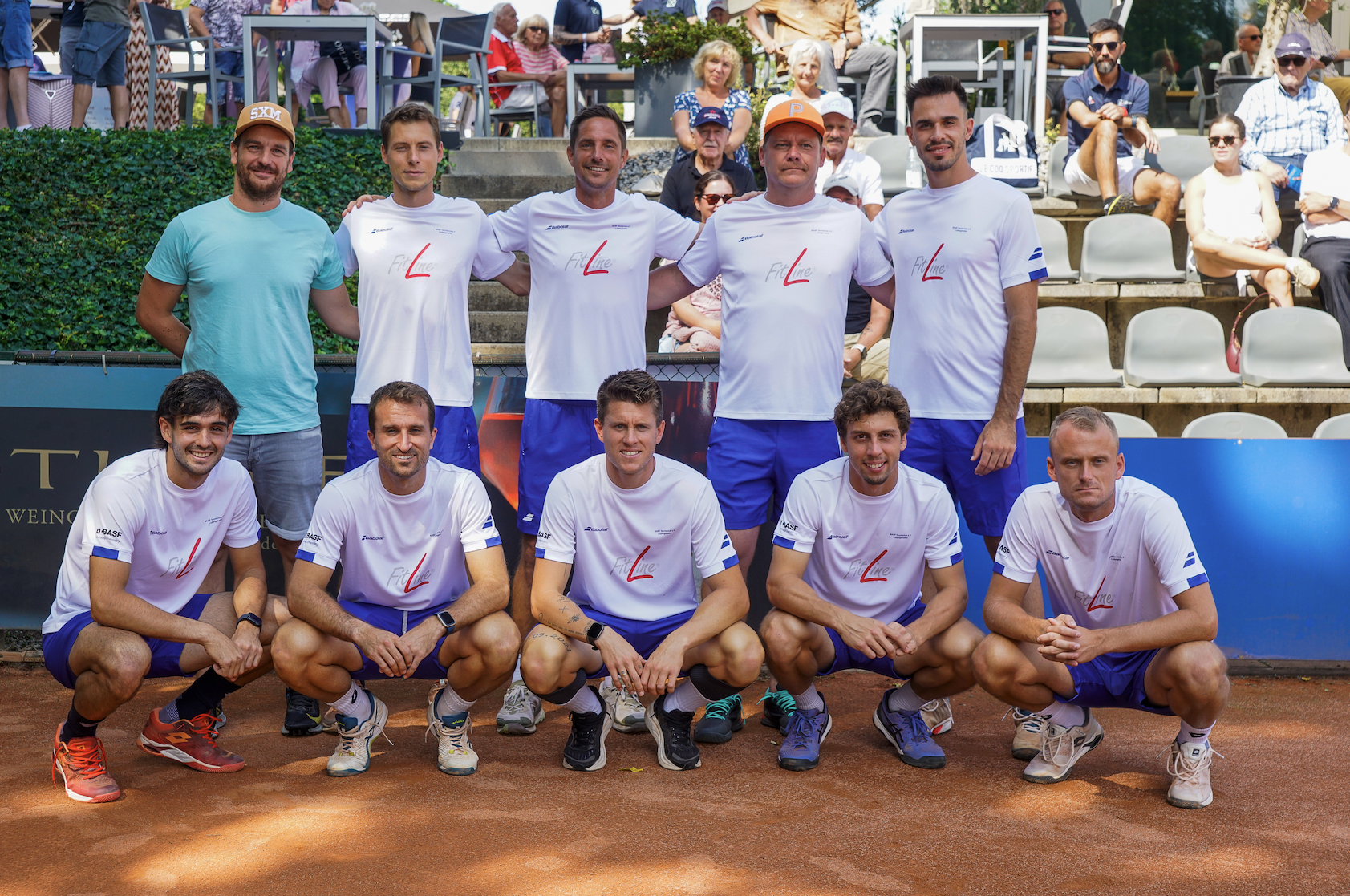 Tennismannschaft posiert auf Sandplatz vor Zuschauern